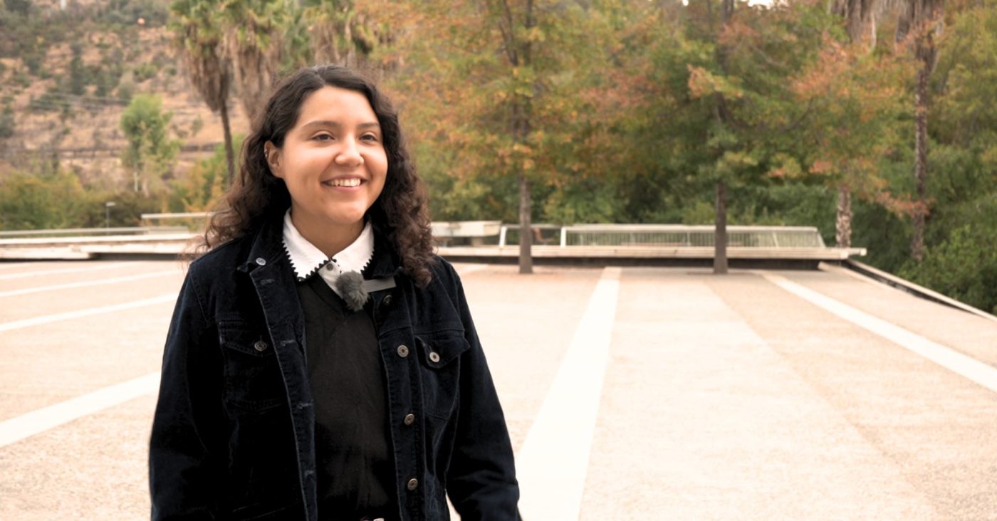Smiling woman with curly hair in a dark jacket standing on a sunlit park path with trees in autumn colors behind her