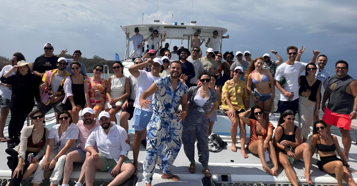 Group of people posing on a sunny boat deck, many in swimsuits with a cabin behind them.
