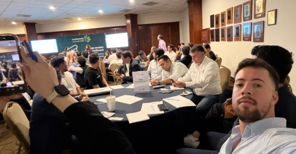 Selfie-style photo of a man in a light shirt at a round-table conference, with attendees working and a panel presenting in the background