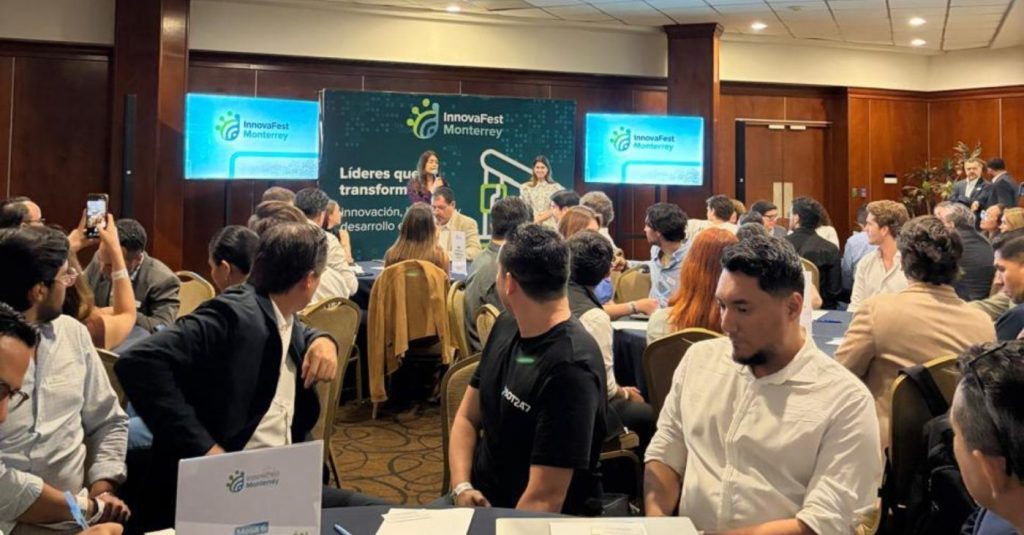 Conference room with round tables and many attendees listening to speakers on a stage with InnovaFest Monterrey banners in the background.
