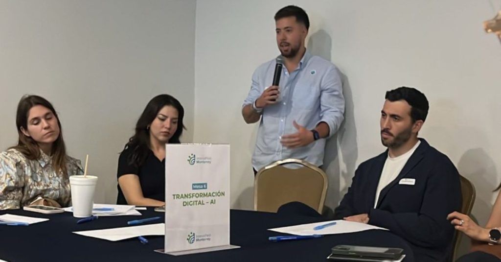 A man stands with a microphone speaking to a panel seated at a round-table about digital transformation and AI, with two women and another man listening.