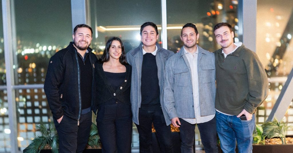 Five friends posing together on a balcony at night with city lights in the background, all smiling.
