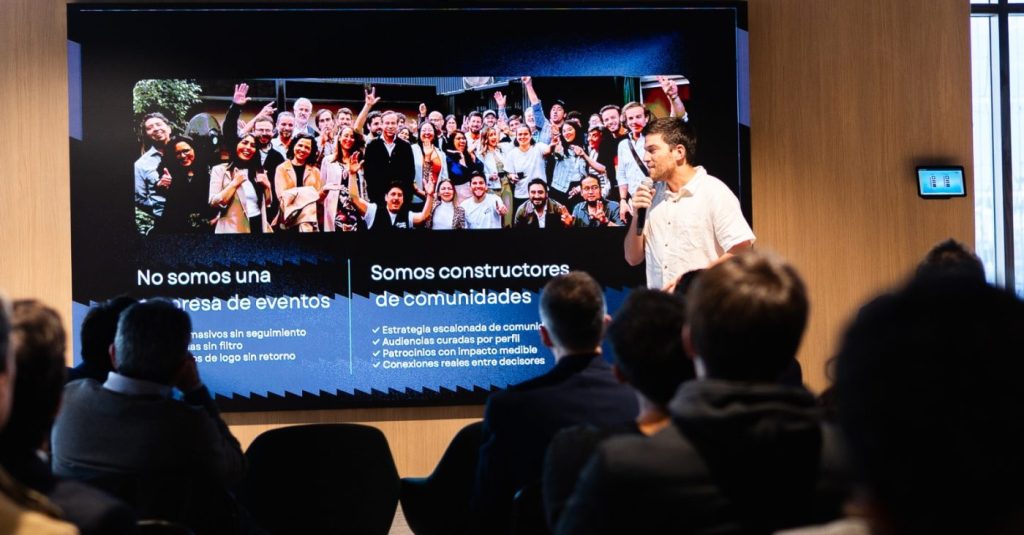 Speaker with a microphone presenting in a conference room; a large screen shows a group photo and Spanish text panels in the background.