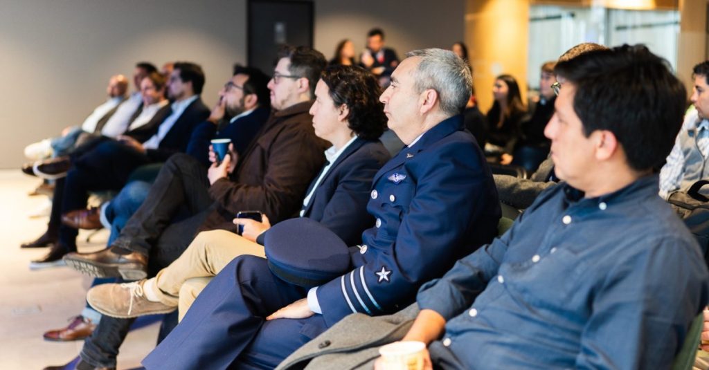 Audience at a conference or seminar, people seated and focused on a speaker; a man in a blue pilot uniform sits near the front with his cap in hand.