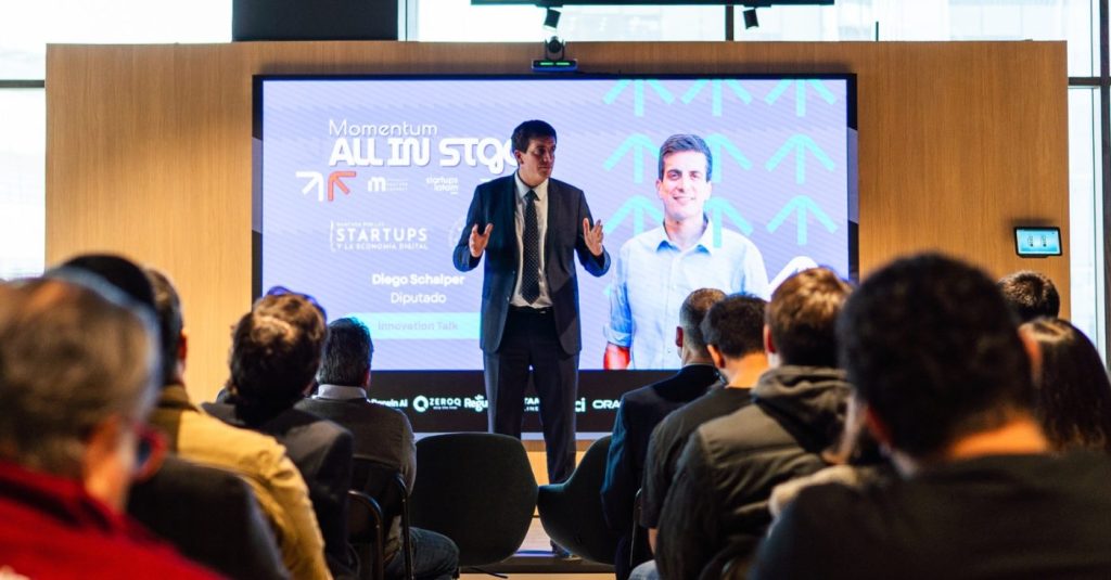 Man in a dark suit giving a presentation on a stage with a large screen behind him at a conference, audience listening attentively.