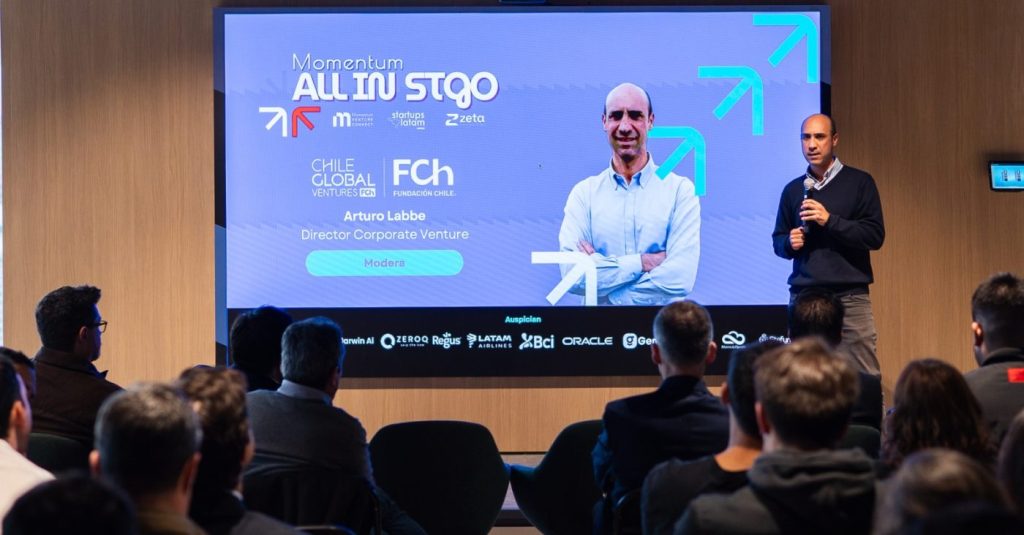 Male presenter on stage holding a microphone, standing beside a large blue slide with logos, addressing a seated audience in a conference room.