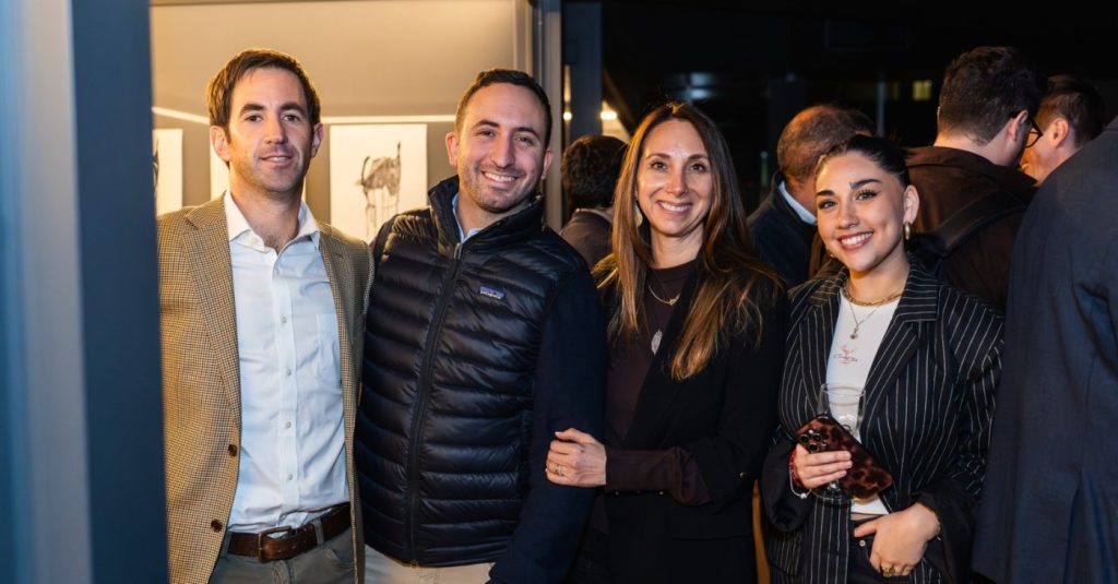 Four adults posing together at a social event, smiling at the camera in smart casual attire.
