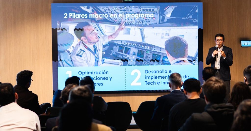 Speaker in a suit presenting to an audience beside a large screen displaying a Spanish slide: '2 Pilares macro en el programa'.