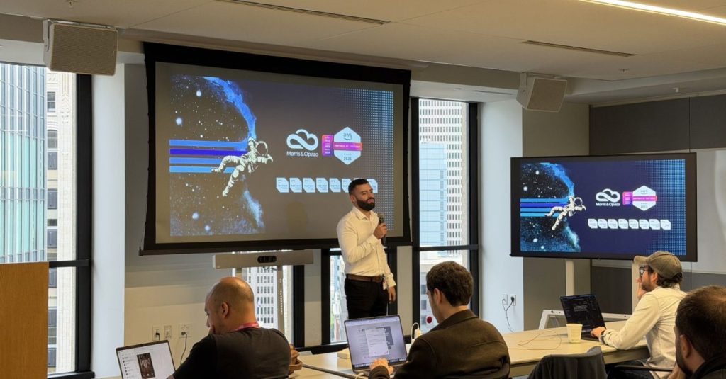 Speaker with microphone presenting in a conference room, two large screens showing a space-themed slide on stage.