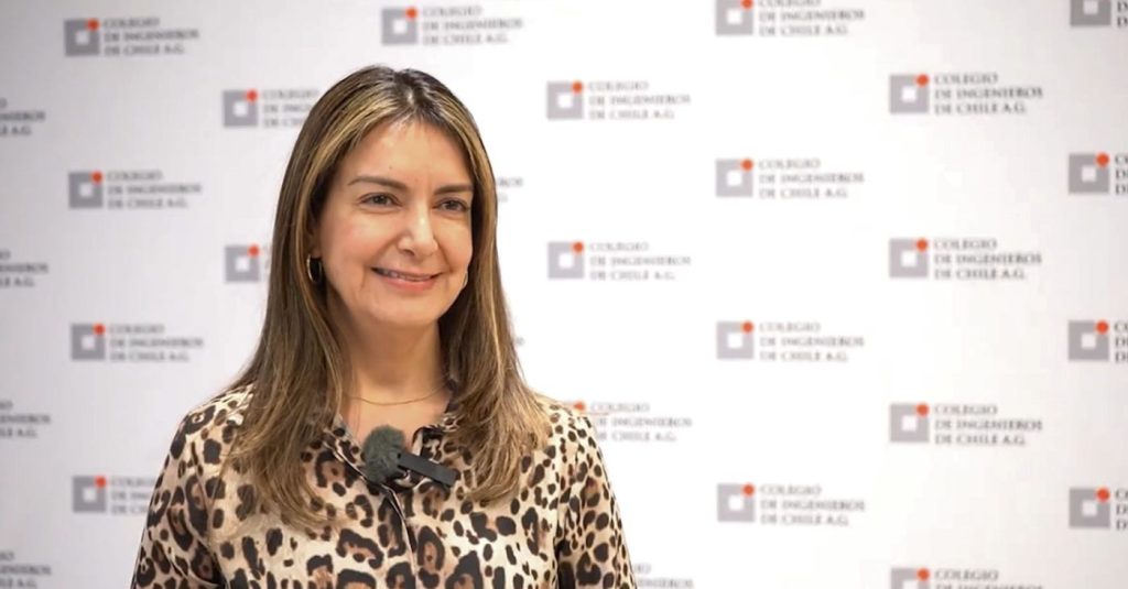 Smiling woman in a leopard-print blouse with a mic clipped to her chest, standing before a backdrop of repeated logos.