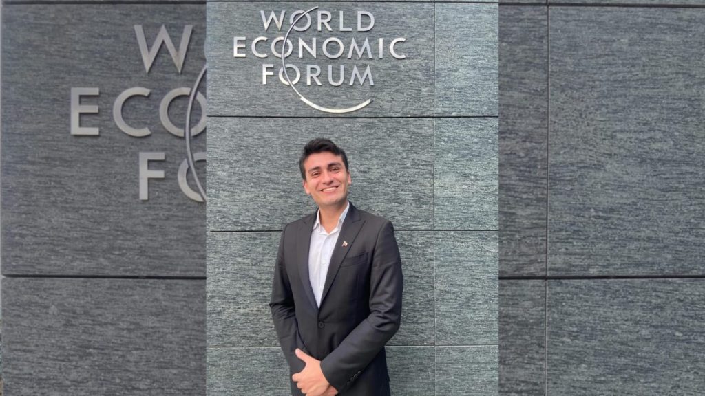 Smiling man in a dark suit standing in front of a World Economic Forum sign mounted on a gray stone wall.