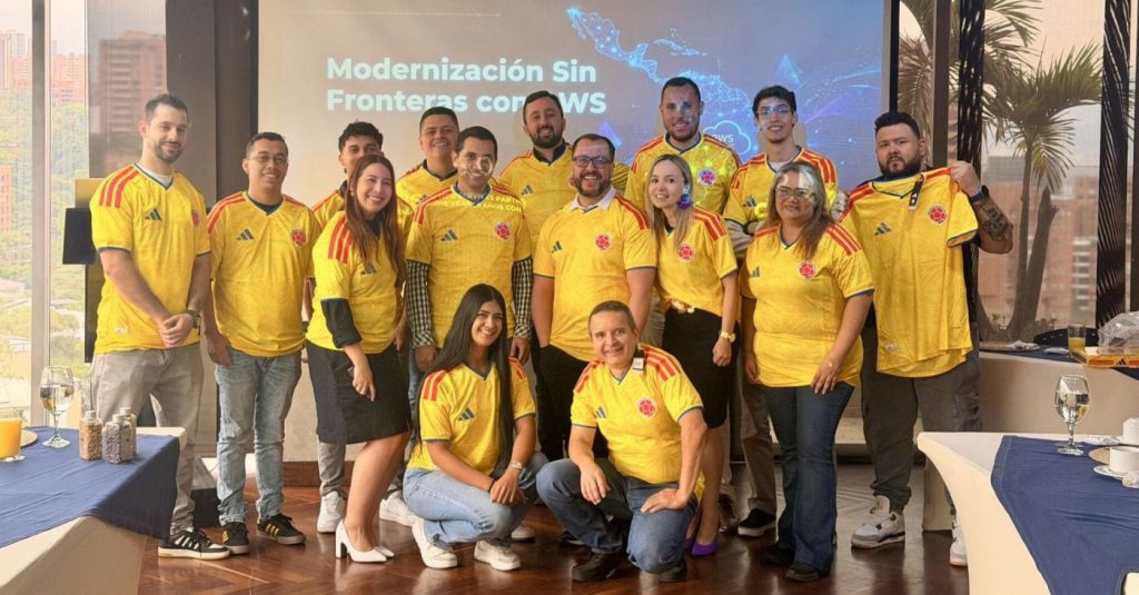 Group of people in yellow Colombia jerseys posing for a group photo at a conference; screen behind says 'Modernización Sin Fronteras con AWS'.','Team photo at a tech event with attendees wearing matching yellow jerseys and smiling.','Mixed group of men and women in yellow jerseys posing indoors for a company event photo.