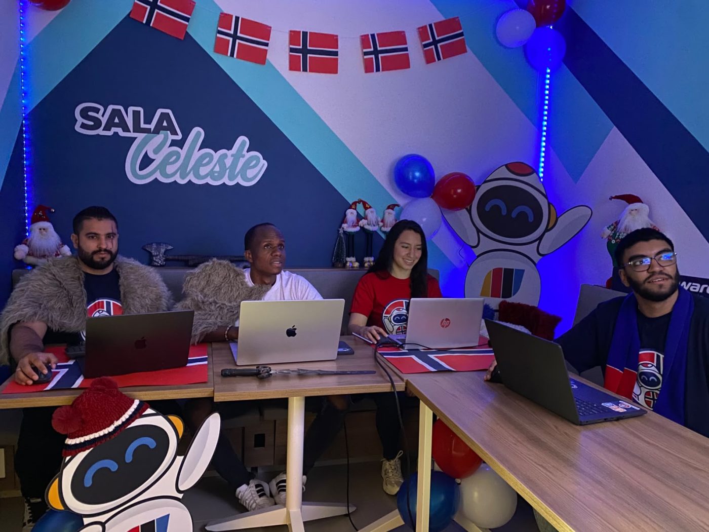 Four people sit at a table with laptops in a festively decorated room, Nordic flag garlands overhead and balloons in blue and red. The wall text reads ‘SALA Celeste’.