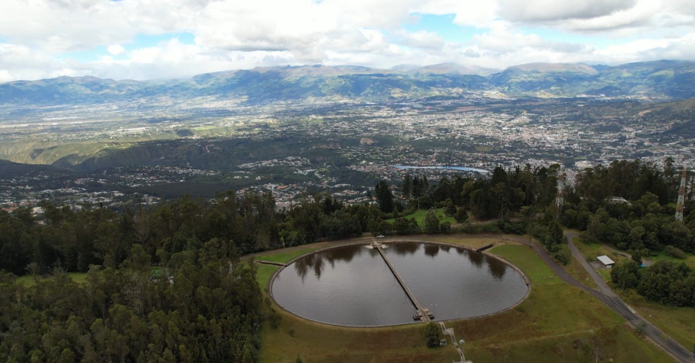 A circular water reservoir on a hillside with a central walkway across the water, overlooking a sprawling city valley and distant mountains.