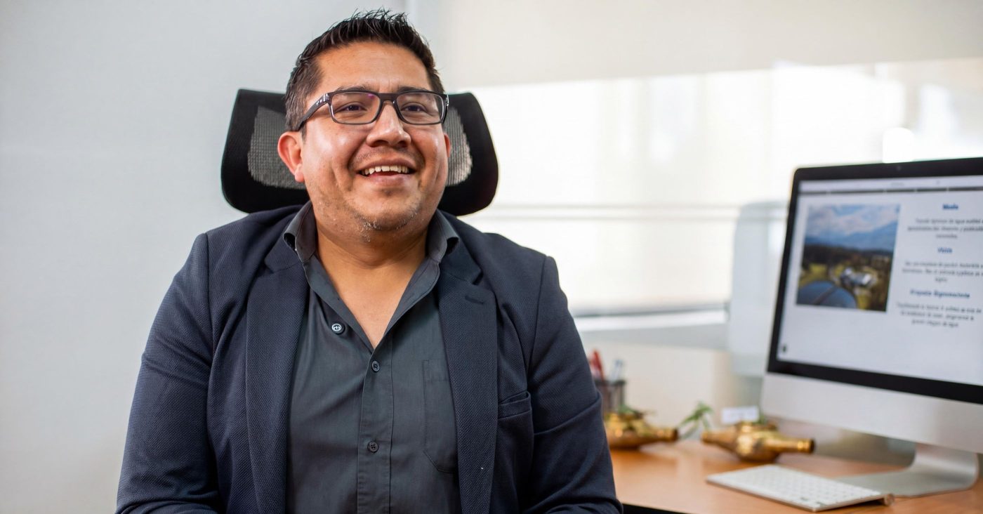 Smiling man wearing glasses and a dark blazer sits at a desk in an office with a computer monitor nearby.
