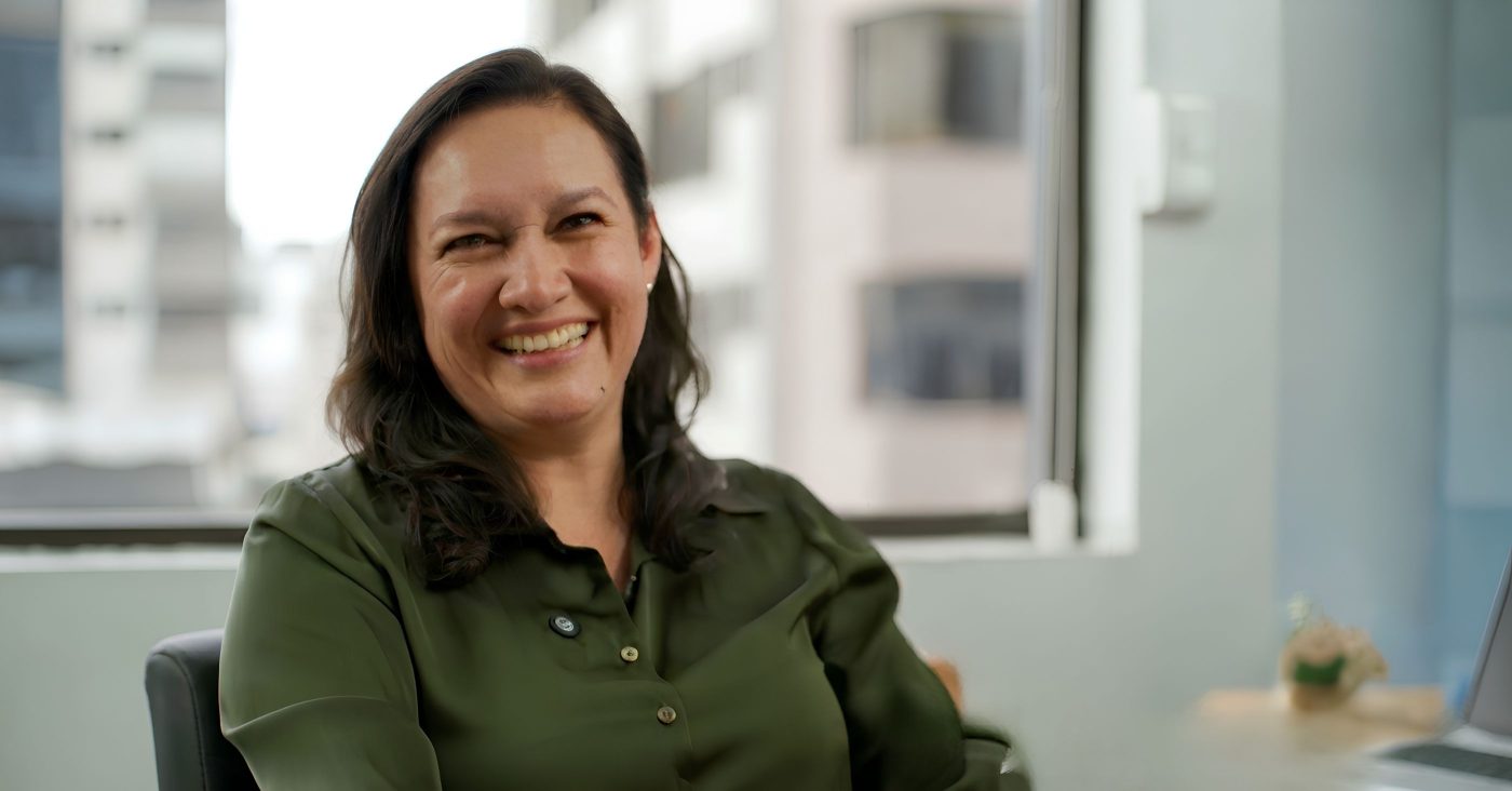 Smiling woman with dark hair in a green blouse sits at an office desk by a window.