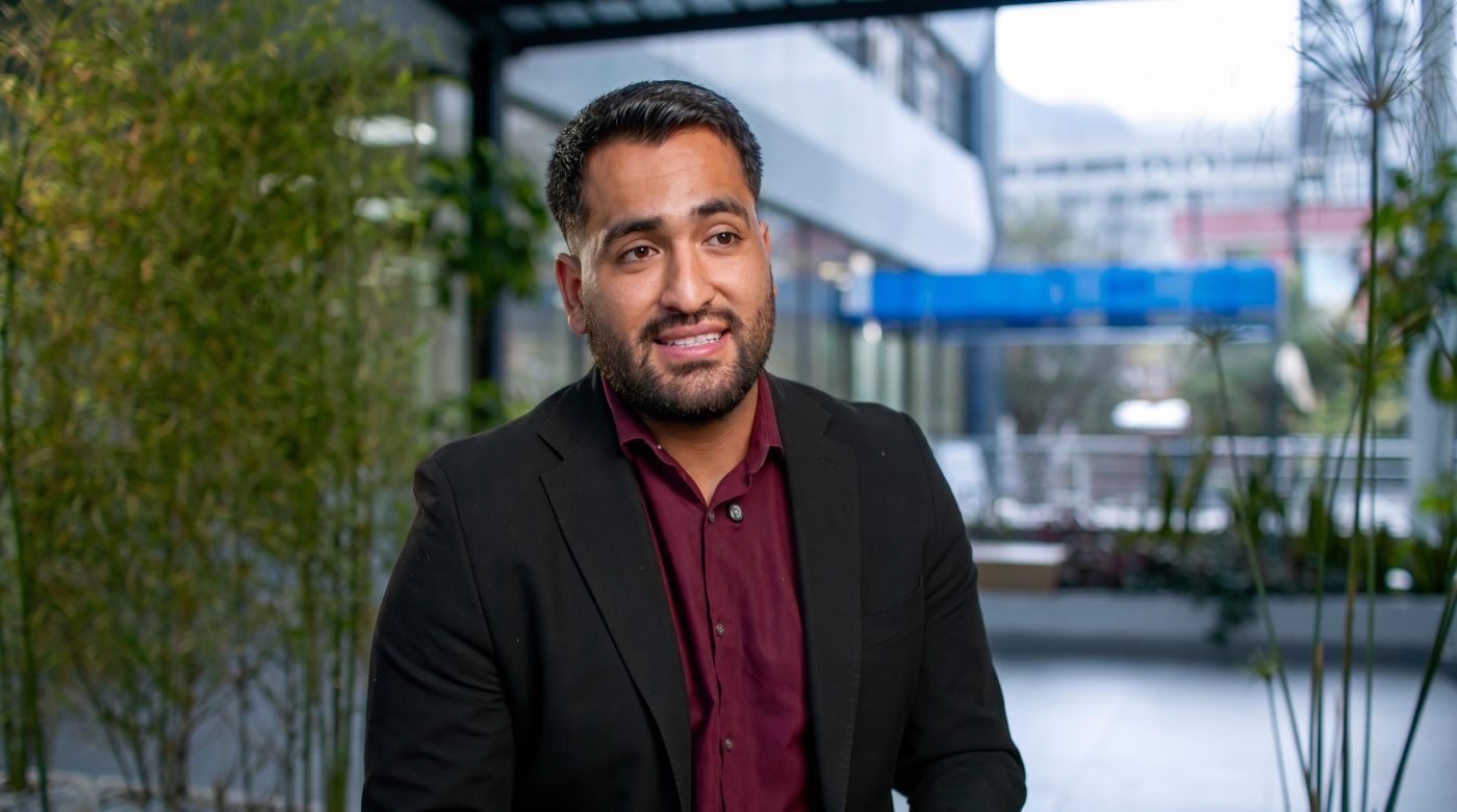 Smiling man in a burgundy shirt and black blazer sits in a modern outdoor courtyard with greenery behind him.